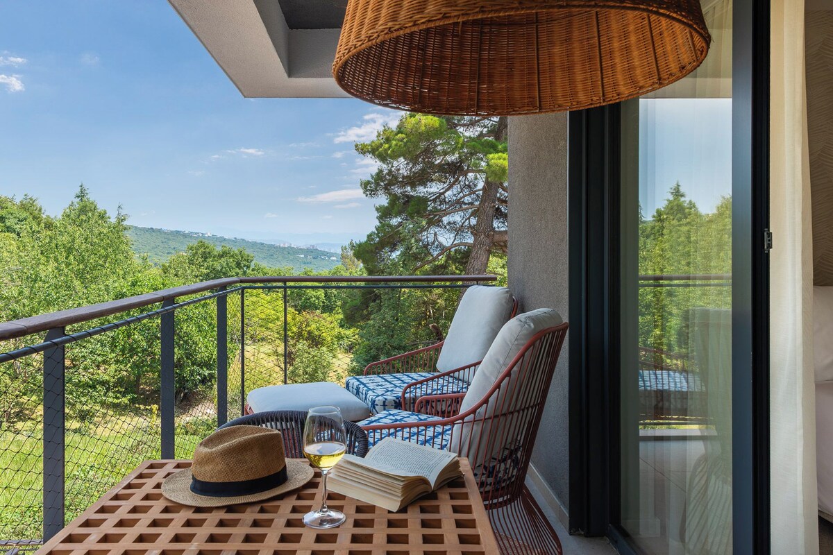 A balcony with wicker furniture, a table with a hat and a wine glass, and a view of greenery and hills.