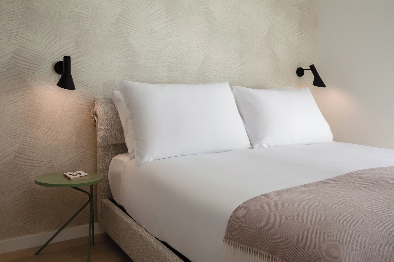 Una cama bien tendida con sábanas blancas, una mesita de noche pequeña verde y dos lámparas fijadas a la pared.
