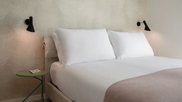 Una cama bien tendida con sábanas blancas, una mesita de noche pequeña verde y dos lámparas fijadas a la pared.