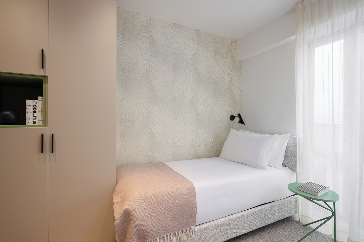 Ein schlicht eingerichtetes Hotelzimmer mit Bett, kleinen Nachttisch, grünem Sessel, Bücherregal und einer Wand mit Muster.