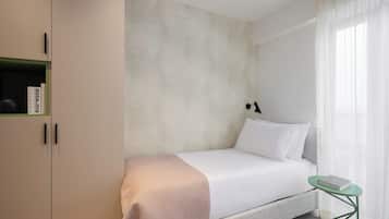 Ein schlicht eingerichtetes Hotelzimmer mit Bett, kleinen Nachttisch, grünem Sessel, Bücherregal und einer Wand mit Muster.