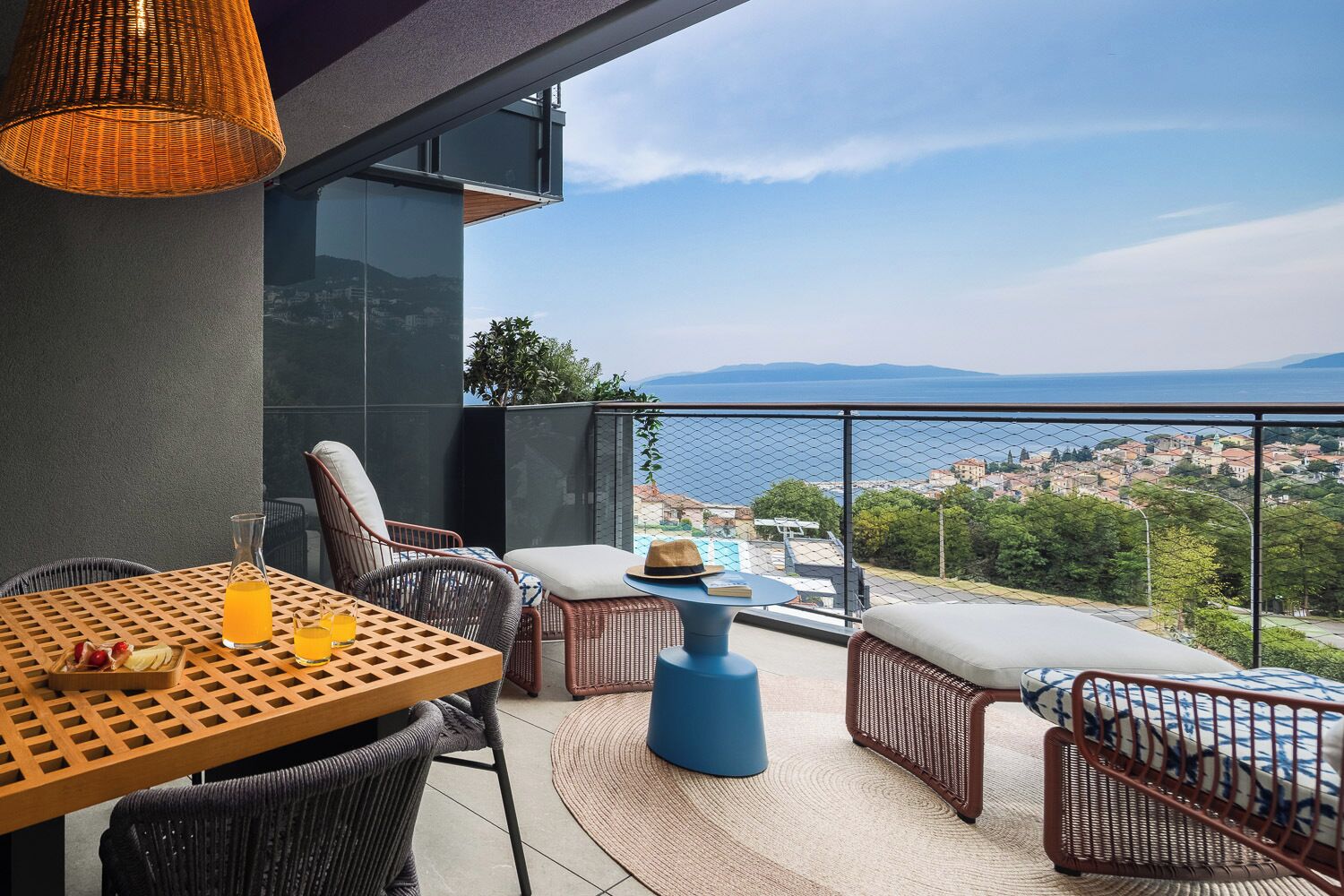 Un balcone con mobili in vimini, un tavolo con bevande e vista su una cittadina costiera.