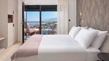 A bedroom with a large bed, a view of a cityscape through a sliding door, and a balcony with outdoor furniture.