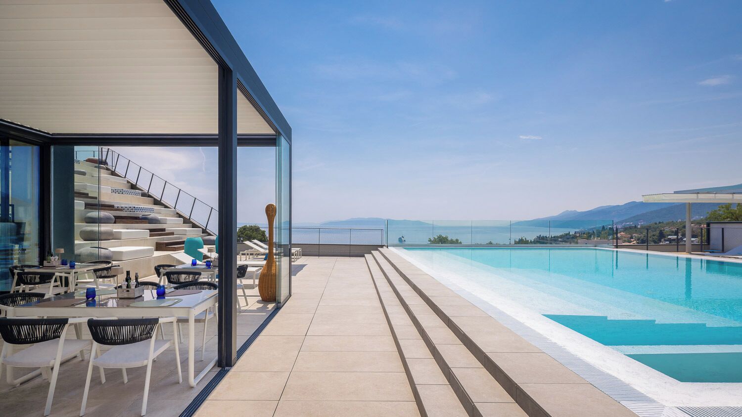 Zona de piscina en la azotea con barandilla de cristal, mobiliario exterior y vista al mar.