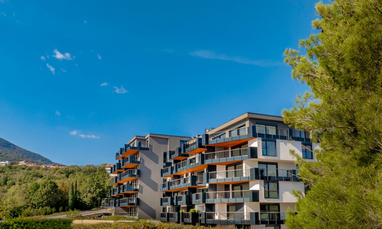 A multi-story residential building with balconies, surrounded by greenery and a clear sky.
