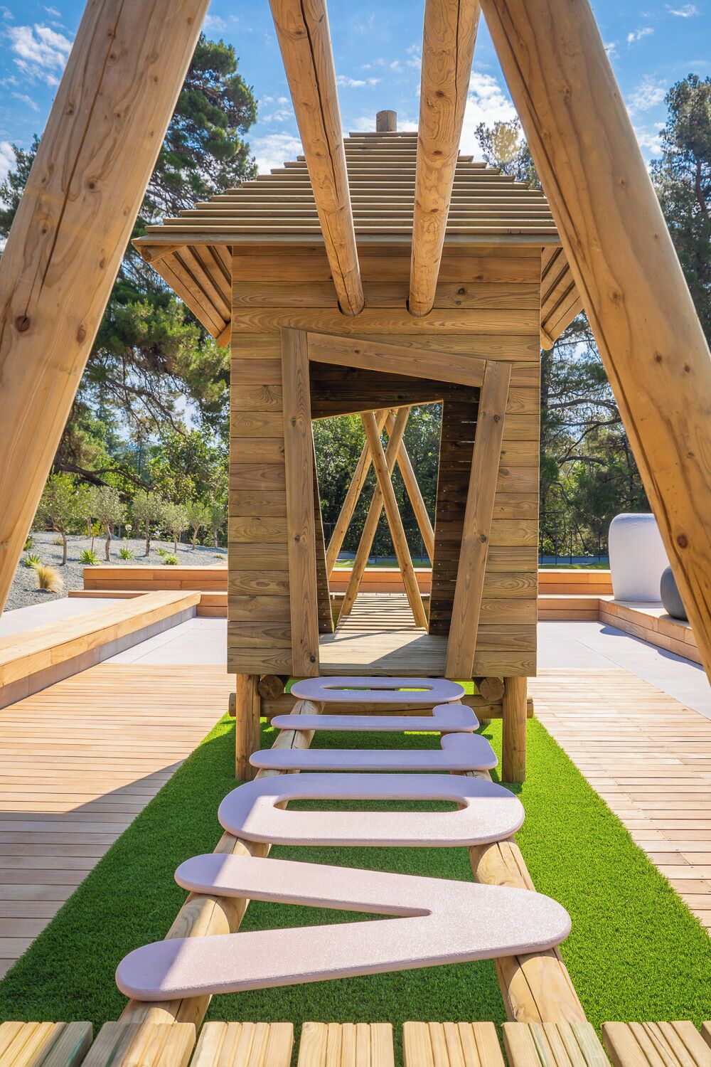 Una estructura de madera con techo inclinado y una abertura central, rodeada por una terraza de madera y césped artificial.