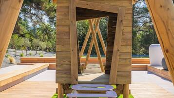 Una estructura de madera con techo inclinado y una abertura central, rodeada por una terraza de madera y césped artificial.