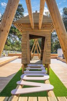 A wooden structure with a sloped roof and a central opening, surrounded by a wooden deck and artificial grass.