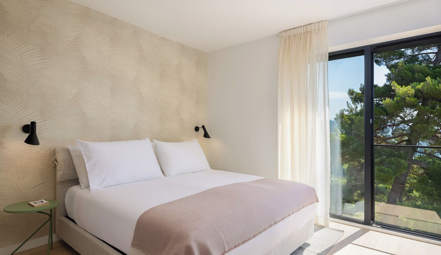 Ein Schlafzimmer mit einem Bett, einem Nachttisch, einem Fenster mit Vorhängen und Blick auf Bäume.
