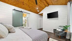 Ein Schlafzimmer mit einem Bett, einer Schiebetür, einem Flachbildfernseher, einem grauen Sessel und einem kleinen Tisch mit einer Pflanze.
