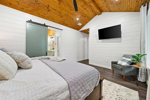 A bedroom with a bed, a sliding door, a flat-screen TV, a gray armchair, and a small table with a plant. - Honey Love Cabin | Hot Tub, Outdoor Fireplace, Near Broken Bow Lake (Broken Bow)