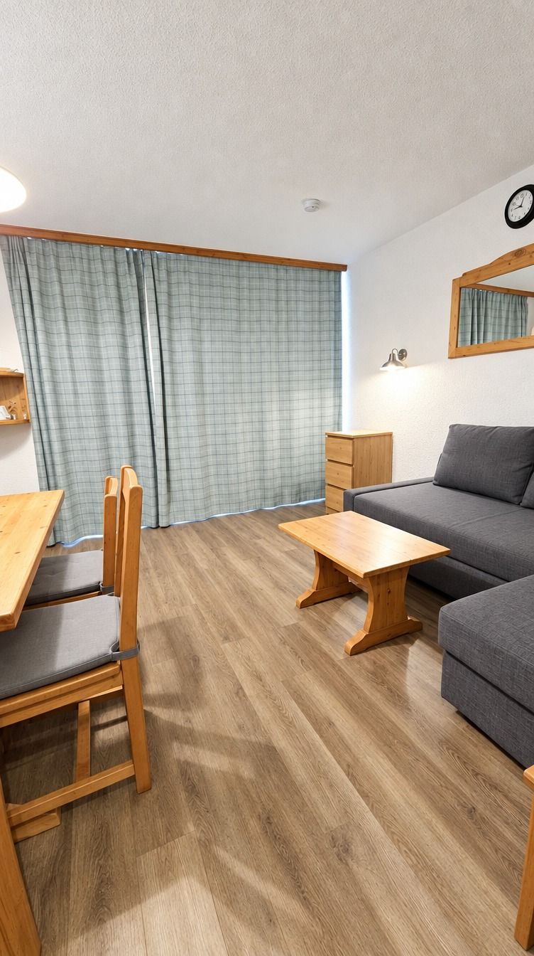 Una habitación con suelo de madera, un sofá gris, una mesa de madera, una silla y un pequeño armario de madera.
