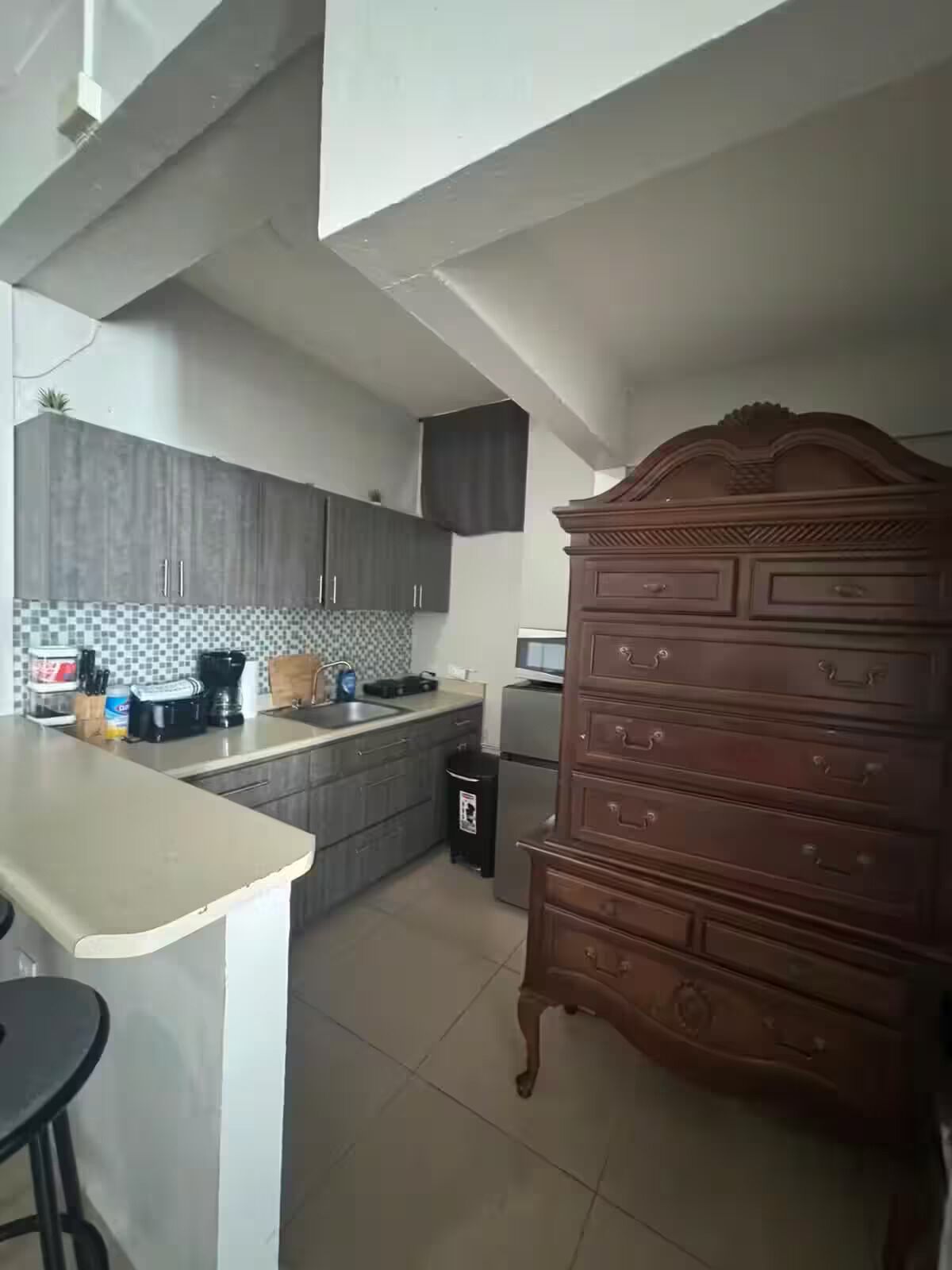 A kitchen with grey cabinets, a wooden dresser, a tiled backsplash, and a small trash bin.