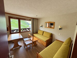 A room with a wooden table, a sofa, a chair, and a window with a view of greenery. - Cozy apartment in the French Alps with direct Ski-In/Ski-Out access. Enjoy stunning mountain views, full comfort after a day on the slopes, and a perfect location close to restaurants and après-ski fun (Les Belleville)