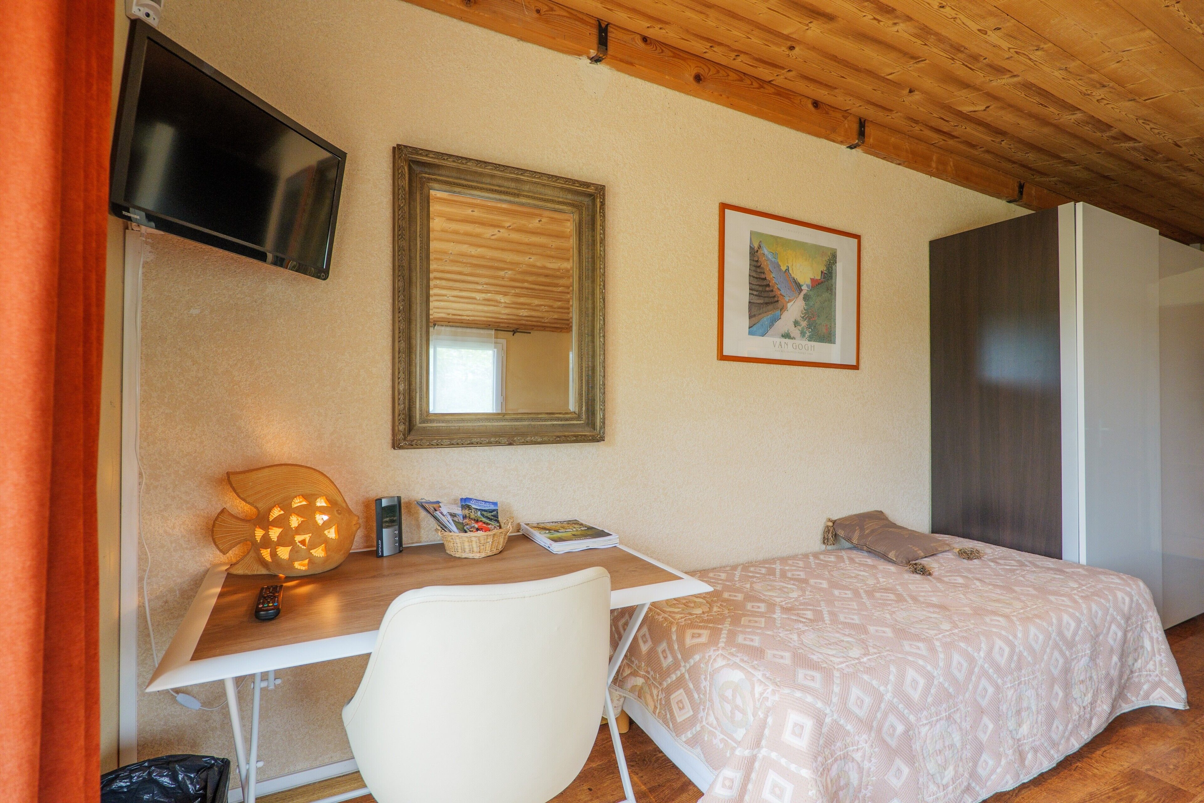 Un dormitorio con cama, escritorio con lámpara, televisión, espejo y un cuadro en la pared.