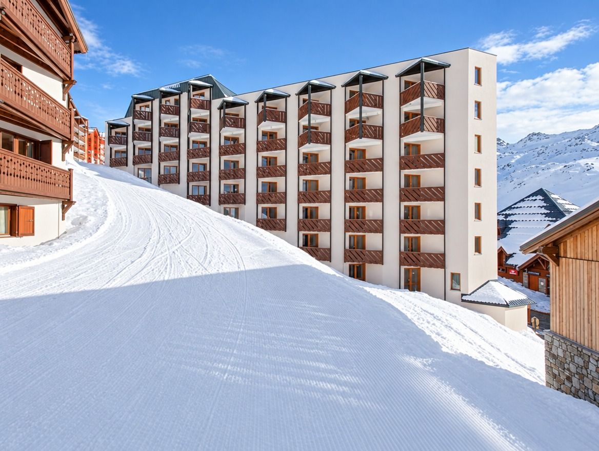 Ein mehrstöckiges Hotel mit Balkonen, eine Skipiste im Vordergrund und eine Berglandschaft im Hintergrund.