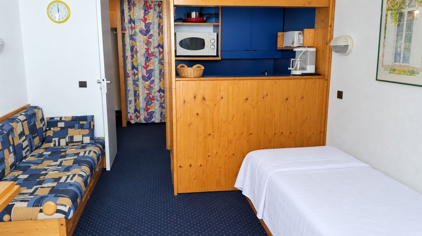 Une pièce de taille modeste comprenant un lit, un canapé, une kitchenette équipée d’un four à micro-ondes et une horloge murale.