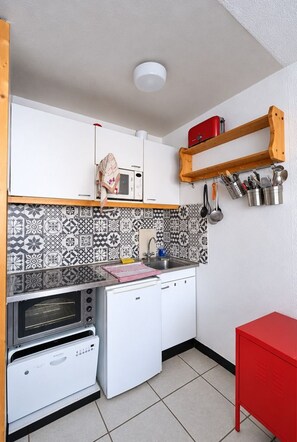 A compact kitchen with a microwave, oven, and dishwasher. There is a wooden shelf above the sink with utensils and a red cabinet to the right. - Val Thorens - Ski In/Out - 6 Beds Flat (N-194) (Les Belleville)