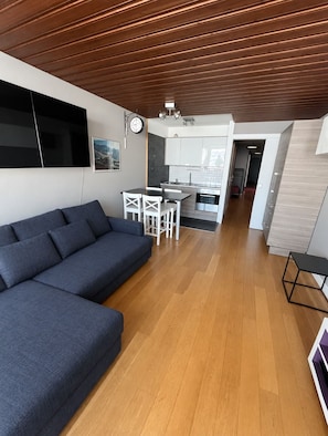 Un espace de vie moderne avec un plafond en bois, un canapé gris, un coin repas avec des chaises blanches et une cuisine en arrière-plan.