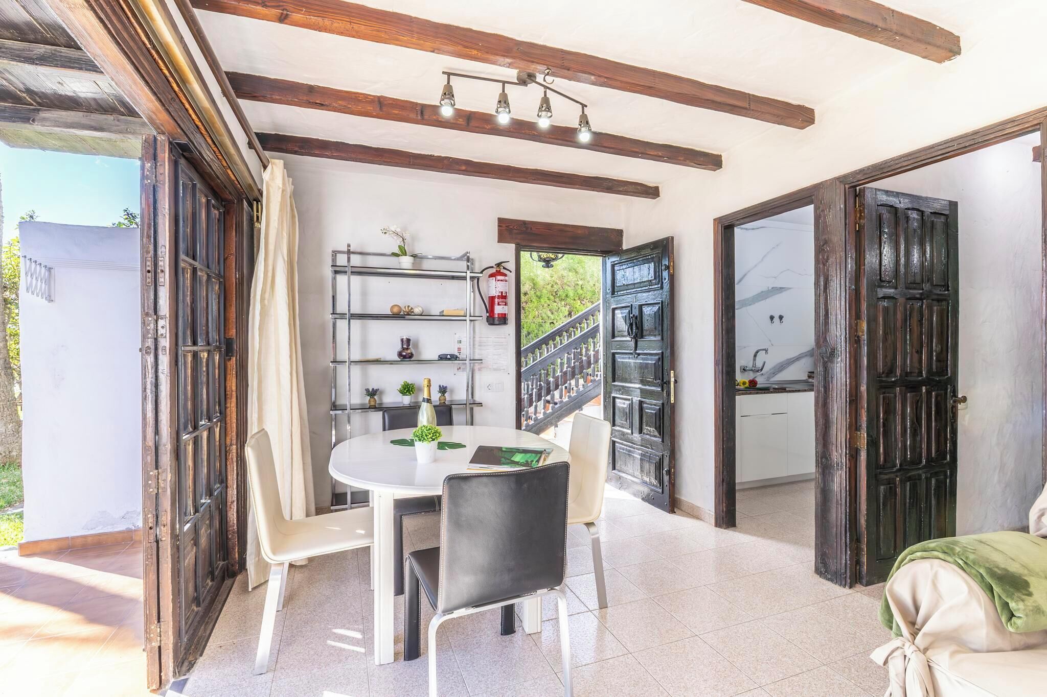 A dining area with a round table, chairs, a shelf with kitchen items, and a view of the outside.