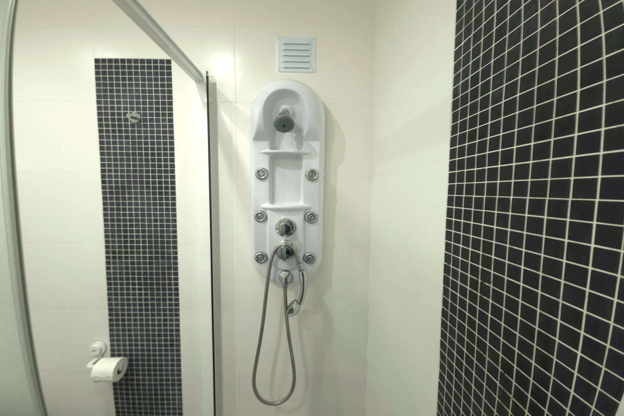 Eine Dusche mit Bedienfeld, Toilettenpapierhalter und Wandlüfter.