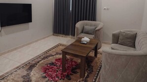 Ein Wohnzimmer mit Flachbildfernseher, zwei beigen Sesseln, einem Holztisch und einem dunklen Vorhang.