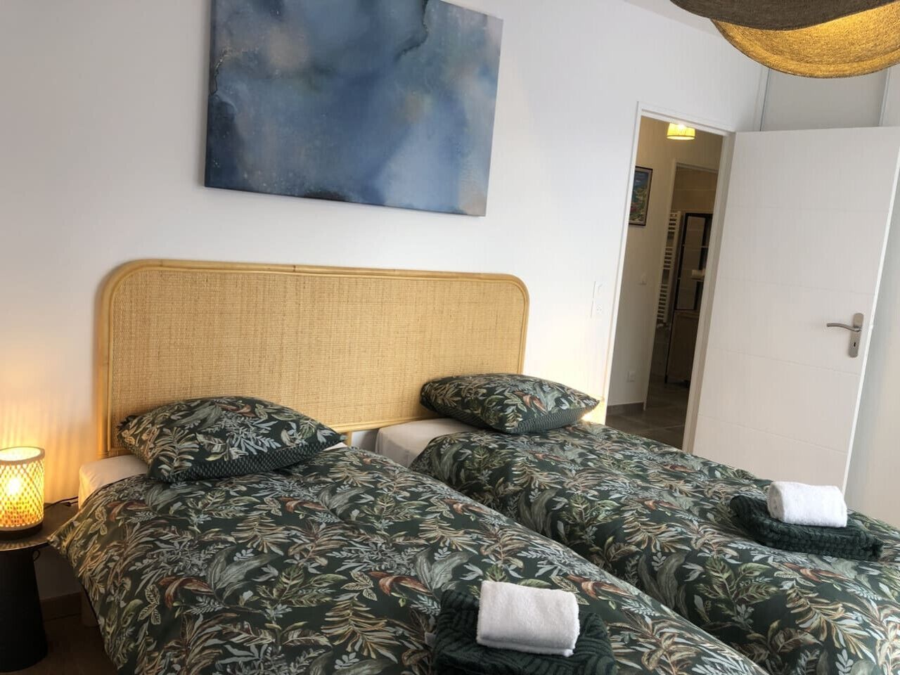 A double room with two beds, a woven headboard, a wall-mounted abstract painting, and a bedside lamp.