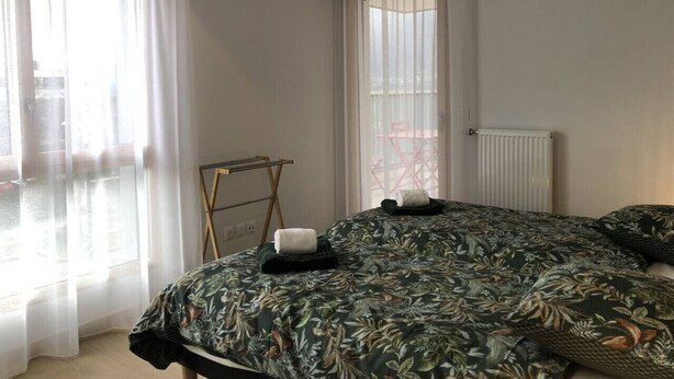 Un dormitorio con cama, lámpara colgante, radiador y ventana con cortinas traslúcidas.
