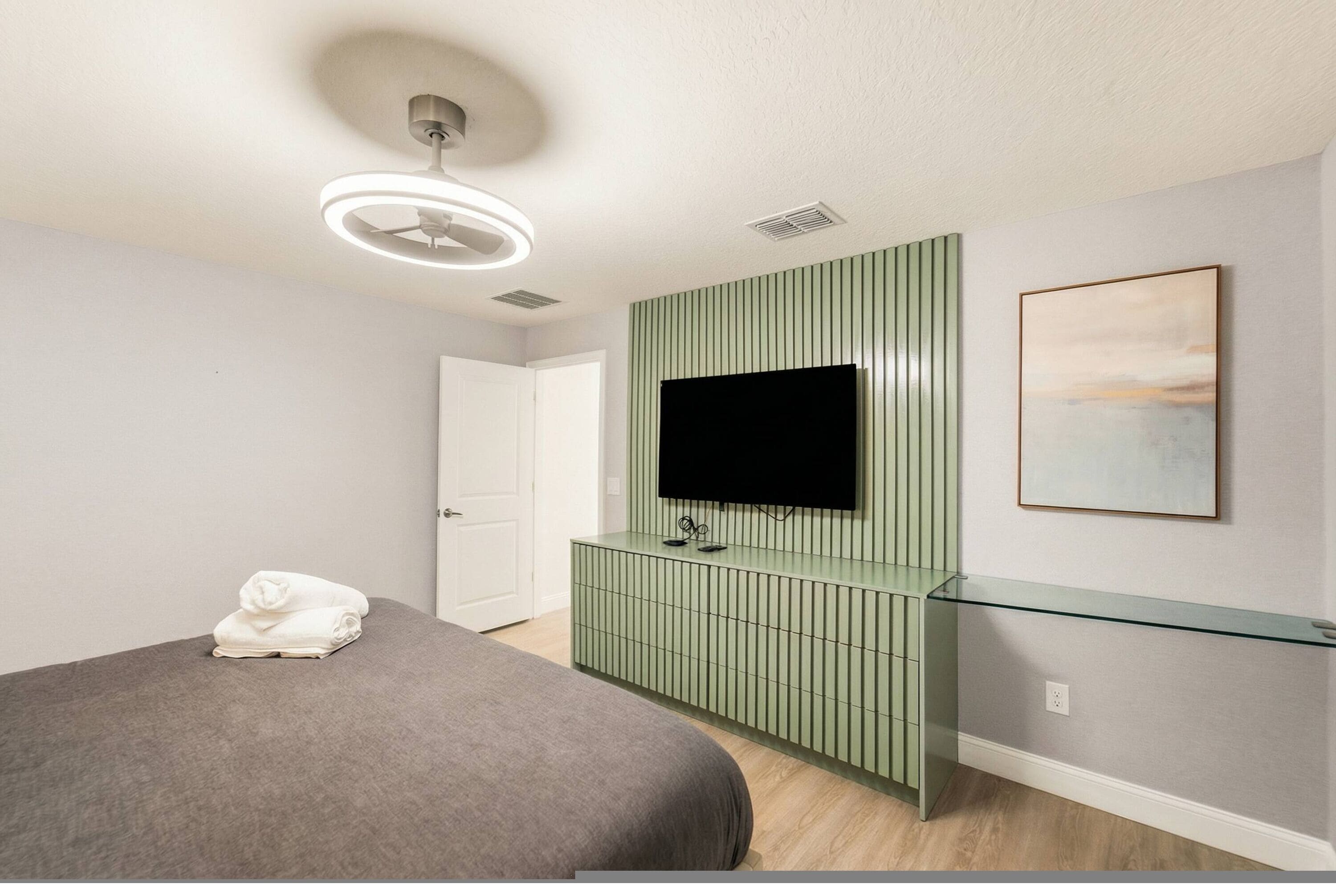 A modern bedroom with a bed, a flat-screen TV mounted on a green panel, a glass console table, and a framed picture on the wall.
