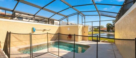 Une piscine extérieure couverte, dotée d’un toit en métal, entourée d’une clôture, et un espace aménagé au bord de la piscine avec un tuyau d’arrosage.