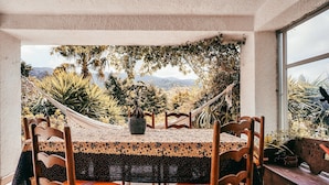 Een eethoek met een tafel voor vier personen, omringd door houten stoelen met oranje kussens, een hangmat en uitzicht op bergen en begroeiing.