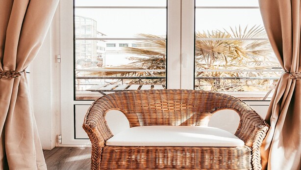 Une chaise en osier avec un coussin blanc, placée devant une fenêtre ornée de rideaux beiges.