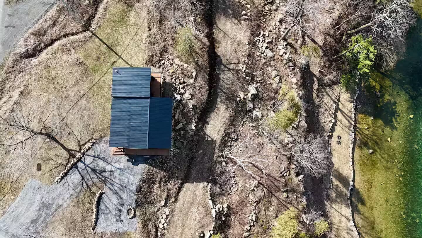 An aerial view of a house with a dark roof, surrounded by rocky terrain and sparse vegetation.