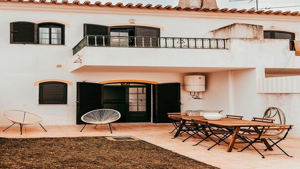Ein zweistöckiges Gebäude mit Balkon, einem Essbereich inklusive Tisch und Stühlen sowie einem kleinen Garten mit zwei Stühlen.
