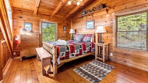 A wooden cabin bedroom with a bed, two bedside tables, a bench, a ceiling fan, and a window with blinds. - Edge of Glory Cabin | Views | Hot Tub | BBQ (Pigeon Forge)
