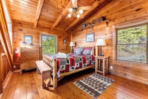 Ein Schlafzimmer einer Holzhütte mit einem Bett, zwei Nachttischen, einer Bank, einem Deckenventilator und einem Fenster mit Jalousien.
