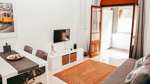 Una sala de estar moderna con un sofá blanco, una mesa de comedor con sillas, un televisor sobre un mueble blanco y un ventanal con cortinas.