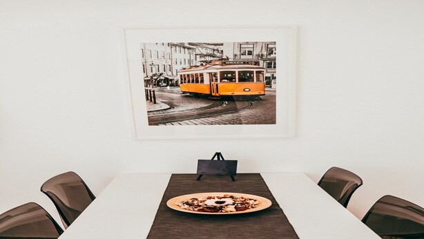 Una mesa de comedor con un plato de comida, un cuadro de un tranvía y un bolso negro sobre un atril.