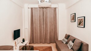Una sala de estar moderna con un sofá, un televisor de pantalla plana, una mesita con una planta y un amplio ventanal con cortinas.