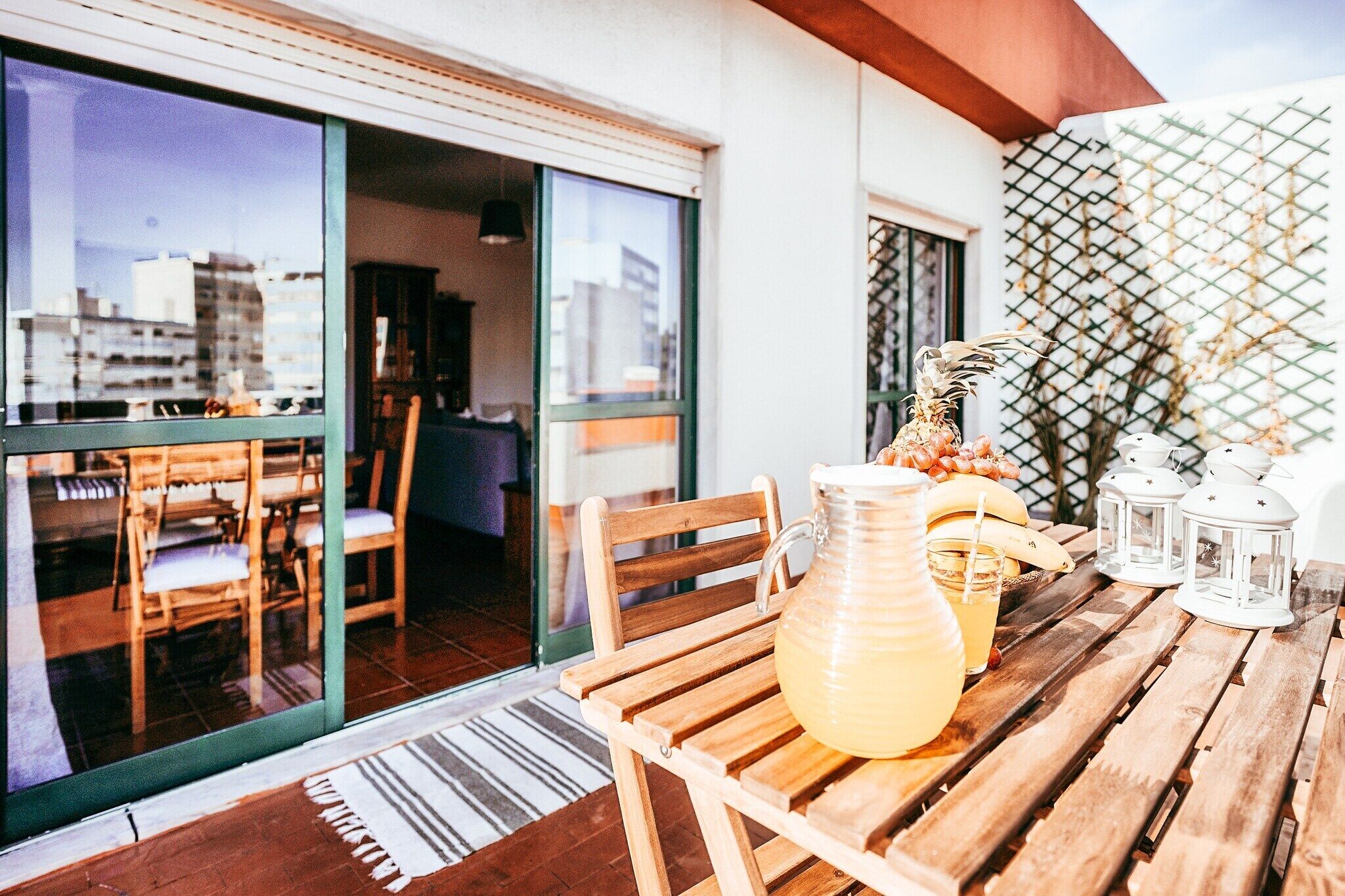 A wooden table with a pitcher of lemonade, a glass, and a banana on a balcony with wooden chairs and a view of buildings.