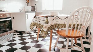 Keittiössä on ruutukuviollinen lattia, valkoinen pöytä, jonka päällä on kukkakuviollinen pöytäliina, ja valkoinen tuoli.