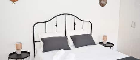 A neatly made bed with white linens and two rolled towels, flanked by two bedside tables with lamps, against a white wall with decorative wall plates.