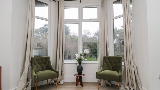 Ein Zimmer mit zwei grünen Sesseln, einem kleinen Holztisch mit Blumenvase und einem großen Fenster mit Vorhängen.