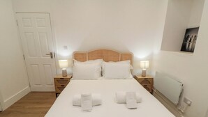 Ein ordentlich bezogenes Bett mit weißen Leinen und zwei Handtüchern, ein Holz-Kopfschild, Nachttischlampen und eine weiße Tür.
