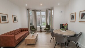 Ein modernes Wohnzimmer mit einem braunen Sofa, zwei grünen Sesseln, einem Holztisch und einem runden Esstisch mit grauen Stühlen.