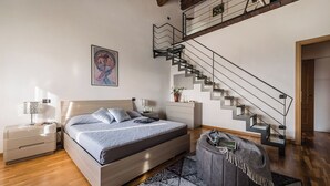 Un dormitorio con cama, mesitas de noche, un taburete gris, alfombra estampada, una escalera y un cuadro en la pared.