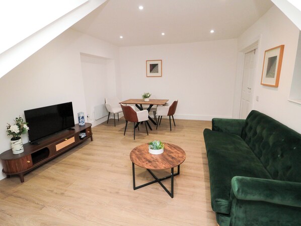 A modern living room with a flat-screen TV, a green sofa, a wooden coffee table, and a dining area with a table and chairs. - Beachwalk Villa 3 (Whitby)