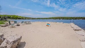 Une plage de sable avec une table de pique-nique et des chaises longues, une petite piscine et une vue sur un lac bordé d’une forêt.