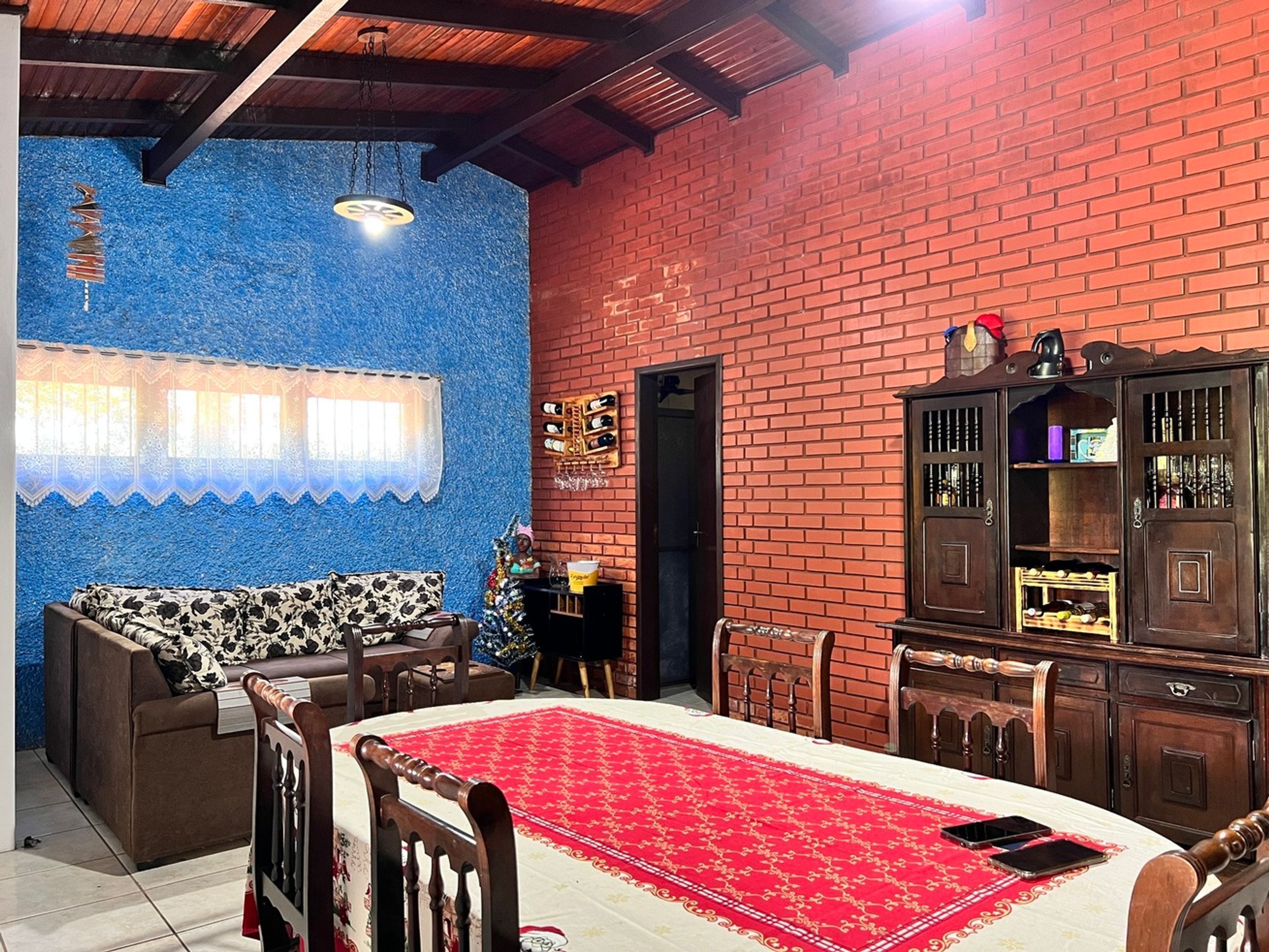 A room with a red brick wall, a blue textured wall, a wooden ceiling, a dining table with a red tablecloth, a sofa, and a wooden cabinet.