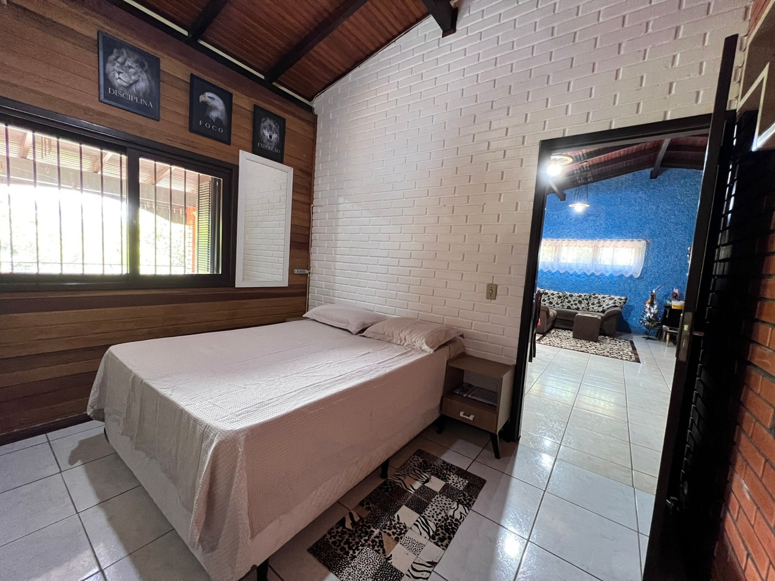 A bedroom with a bed, a window, a white brick wall, and a door leading to a living area.